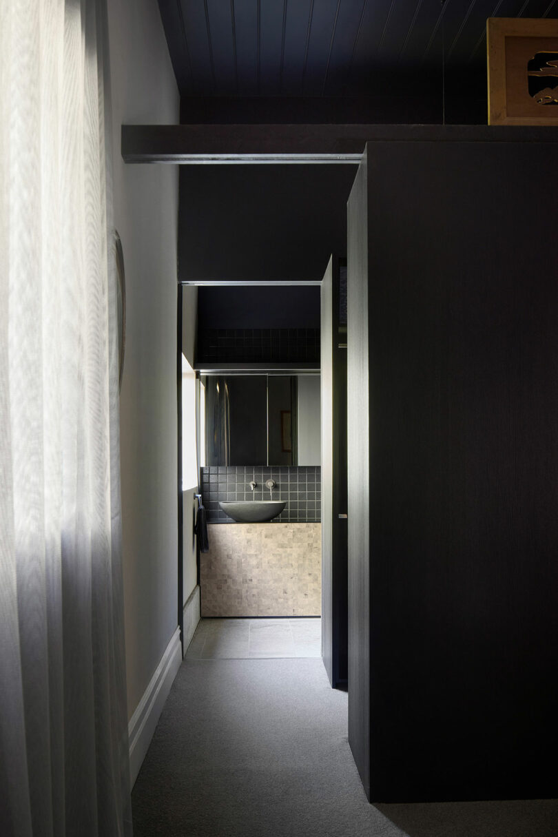 A narrow corridor with dark walls leads to a small bathroom featuring a sink, mirror, and tiled wall. Curtains hang to the left, and light filters in from a window.