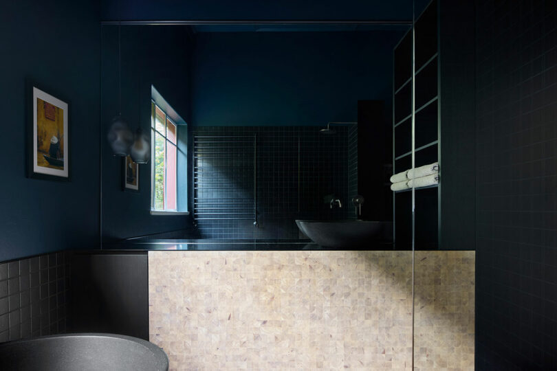 A modern bathroom with dark tiles, a window, a mounted towel rack, a wall mirror, and minimalist fixtures. A framed picture hangs on the blue wall.