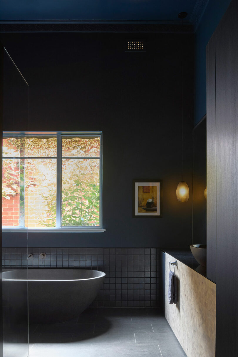 A dimly lit modern bathroom with a dark freestanding bathtub, a wall-mounted washbasin, a large window with leafy view, and hanging pendant lights.