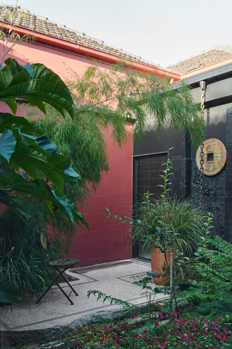 A small courtyard garden features potted plants and greenery against a red and black wall, with a small table and chair set. A roof with tiles is visible above the walls.