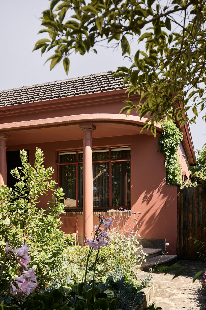 A red house with a tiled roof, porch supported by two columns, and surrounded by lush green and flowering plants, partially shaded by tree branches.