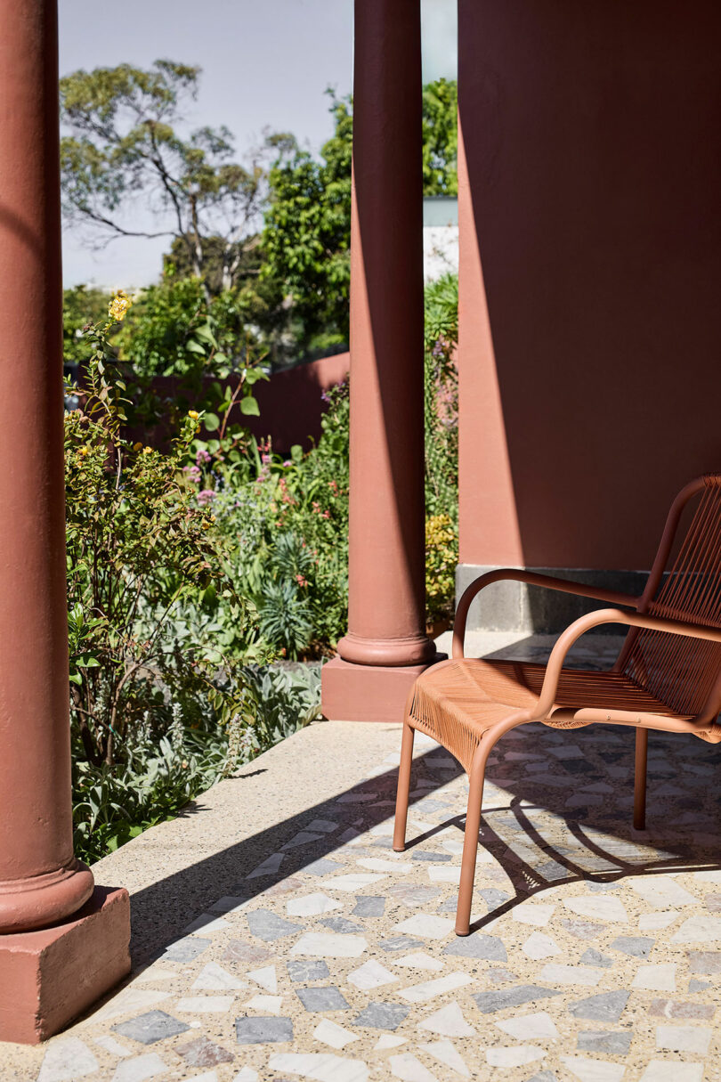 Brick red pillars and a brown chair on a tiled porch overlook a garden with various green plants and trees in the background.