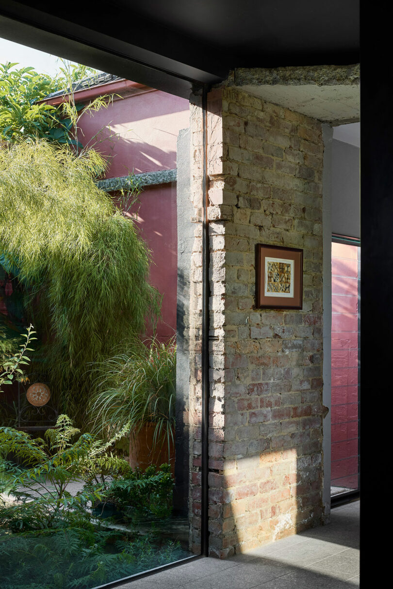 A view of a garden with lush green plants from inside a room through a glass wall, with a brick pillar and a painting hanging on it.