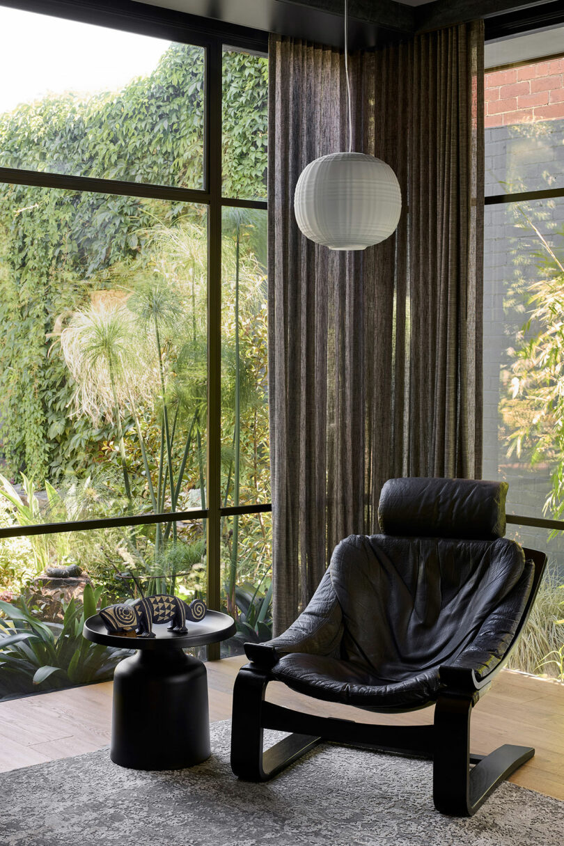 A modern seating area with a black leather chair, a small round table, and a paper lantern light in front of large windows showcasing a lush green garden.