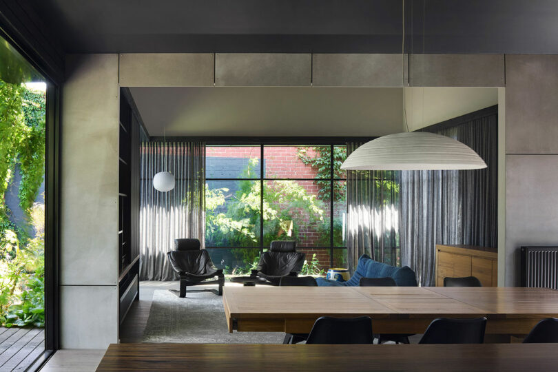 Modern living and dining area with large windows, gray curtains, black armchairs, a blue sofa, a wooden dining table with black chairs, and a large pendant light. Lush greenery is visible outside.