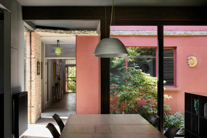 A modern dining area with a large wooden table, chairs, and a hanging light. There's an open doorway leading to a hallway, and a window showcasing an outdoor garden with greenery.