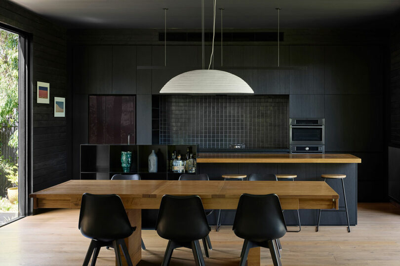 Modern kitchen with dark-toned cabinets, wooden dining table, black chairs, hanging light fixture, and a minimalist backsplash, surrounded by black walls with artwork.