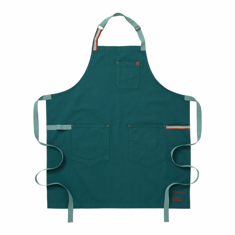 A forest green apron with light green straps and three front pockets is laid flat. The apron has an adjustable neck strap and waist ties, with a small red embroidered label near the bottom right pocket.