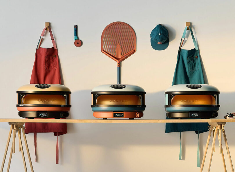 Pizza ovens on a wooden table, with three aprons, a pizza peel, and a cap hanging on the wall behind.