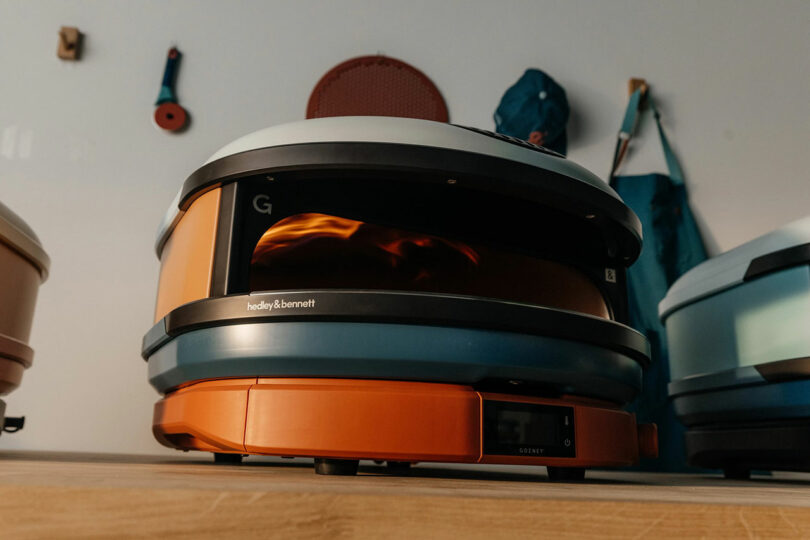 A close-up of an orange and blue tabletop pizza oven with a digital display, showing flames inside. Various items hang on the wall in the background.