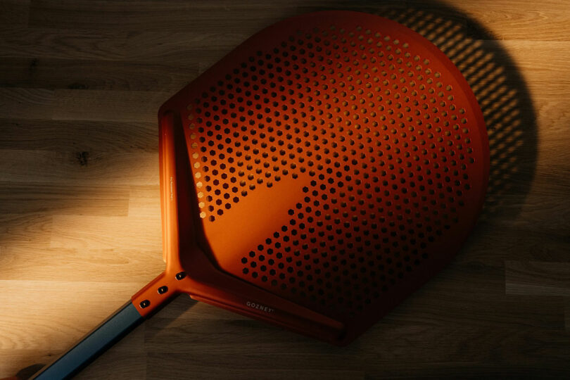 A red perforated pizza peel rests on a wooden surface with light casting shadows through the holes.