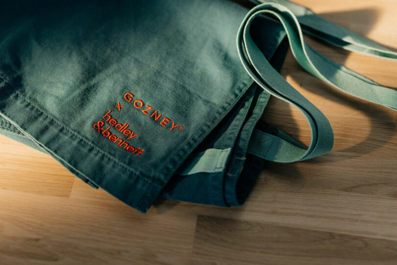 Close-up of a green apron with embroidered text reading "GOZNEY x hedley & bennett" in orange, lying on a wooden surface.