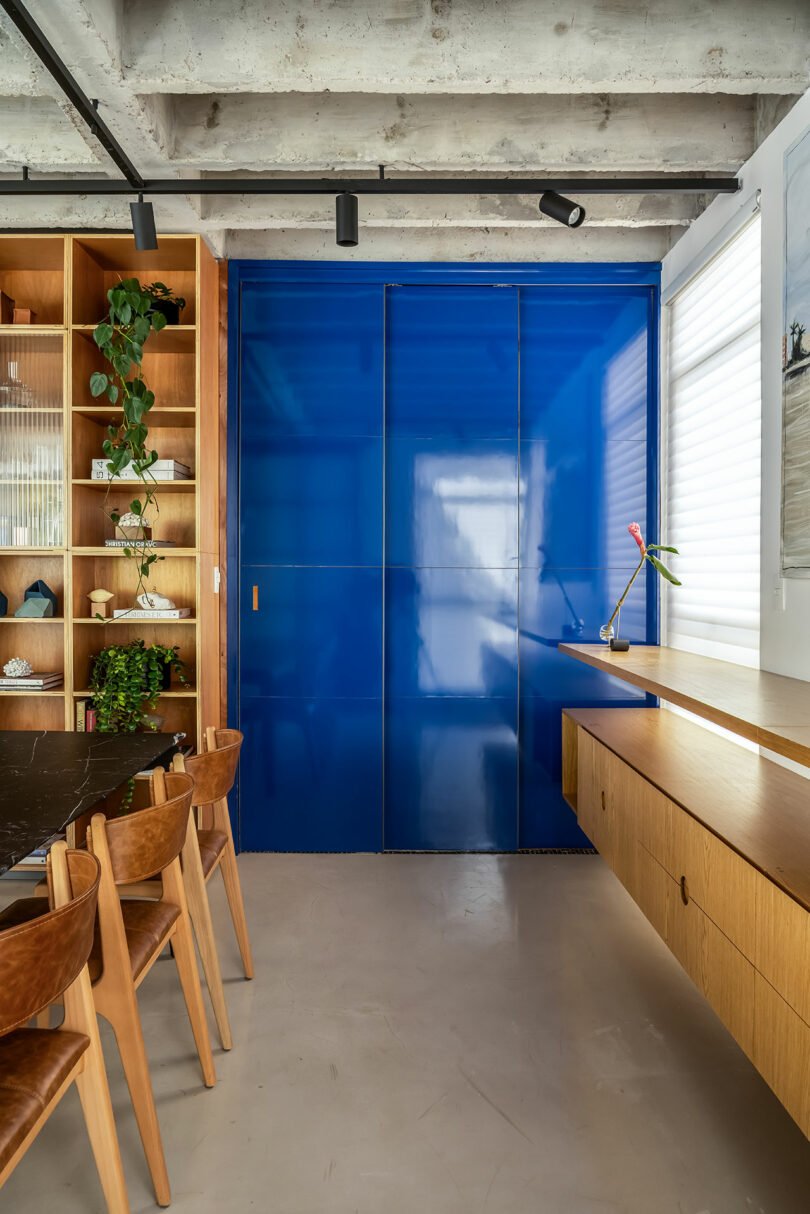 A modern room features a blue sliding door, wooden chairs, a dining table, shelves with plants, and a wooden floating desk under a window. Exposed concrete ceiling above.