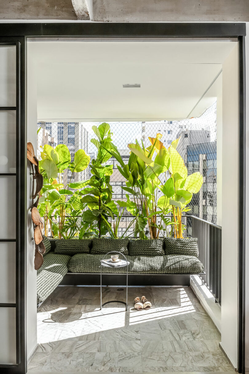 A cozy balcony with a cushioned seating area, a small table, potted plants, and a cityscape view in the background.