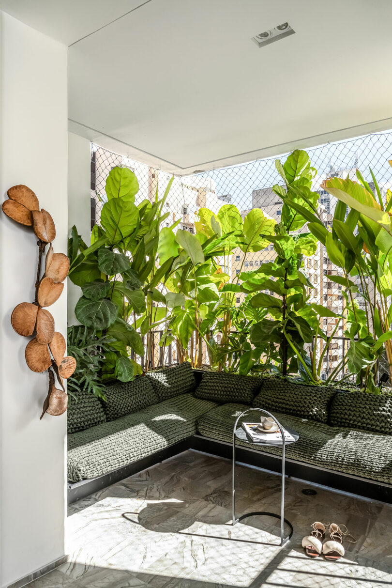 Contemporary indoor seating area with green cushioned bench, surrounded by tall, lush plants, and a small table holding magazines. Sunlight illuminates the area, casting shadows on the tiled floor.