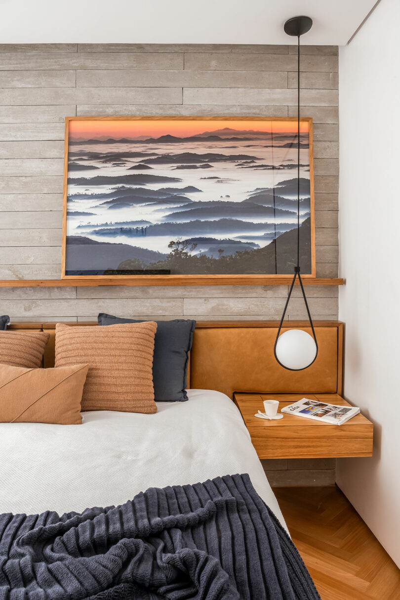 Modern bedroom with a wooden headboard, a mountain landscape painting, a hanging pendant light, and a bedside table with a cup and a magazine. Blue and brown tones dominate the decor.