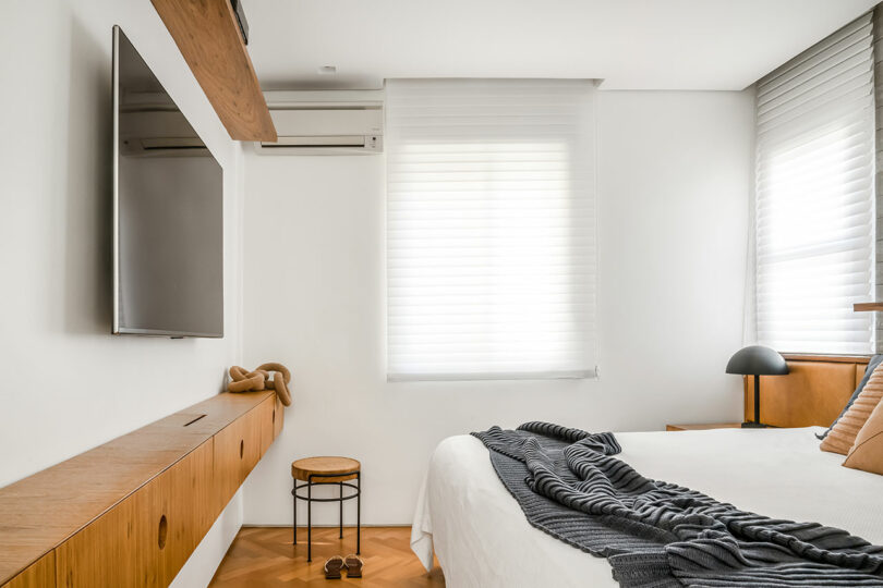 Modern bedroom with a neatly made bed, striped throw, wall-mounted TV, air conditioner, small window-side stool, and minimalistic wooden furniture. Natural light through partially covered windows illuminates the room.