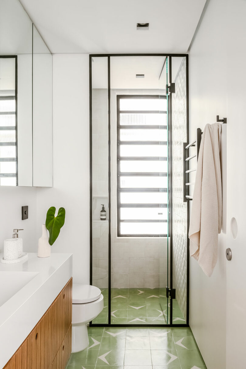 A modern bathroom featuring a glass-enclosed shower, green geometric floor tiles, a wall-mounted towel, and a vanity with a plant and toiletries.