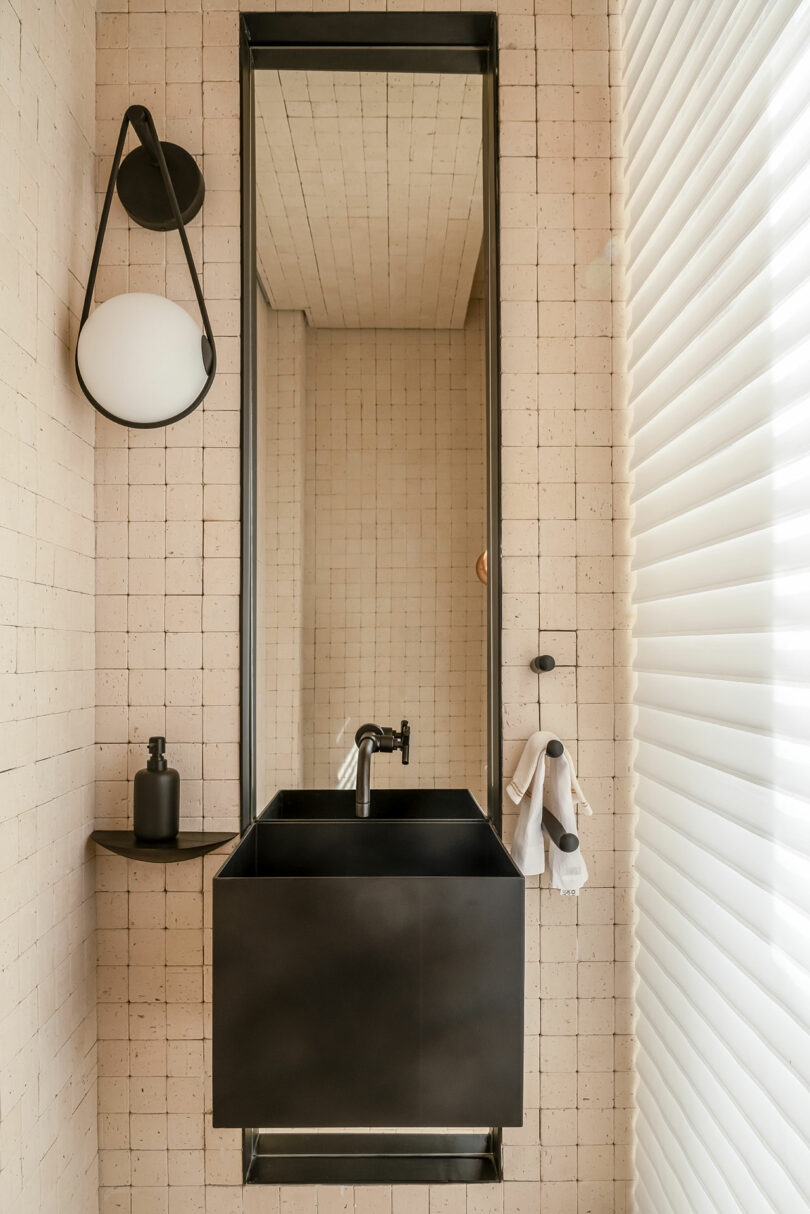 A compact bathroom with a black sink, tall mirror, wall-mounted light fixture, soap dispenser, and a towel hanging beside Venetian blinds on the right. The walls are tiled in a beige grid pattern.