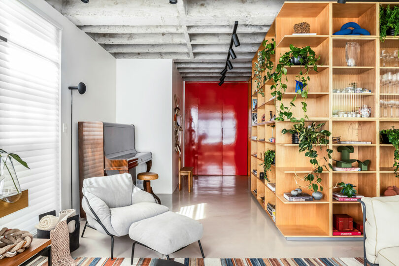 A modern living room features a lounge chair, a wall-mounted piano, a wooden shelving unit with plants and decor, and a red door in the background. The ceiling shows exposed beams.