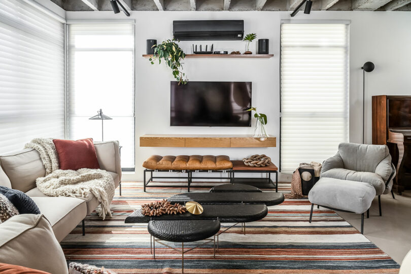 Modern living room with white walls, striped rug, and a wall-mounted TV. Features a beige sectional sofa, armchair, leather bench, and round coffee tables. Indoor plants and decor items are present.