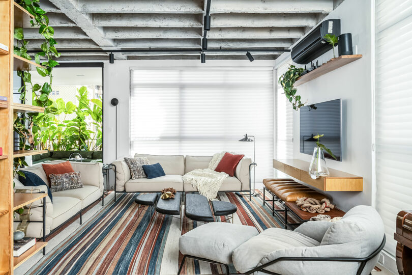 A bright living room with modern furniture, including sofas, a chaise lounge, a striped rug, and shelves with plants. The ceiling has exposed beams, and a large window provides natural light.