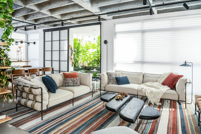 A modern living room features beige sofas with colorful cushions, a striped rug, a coffee table, and large windows with blinds. There are plants and a dining area in the background.