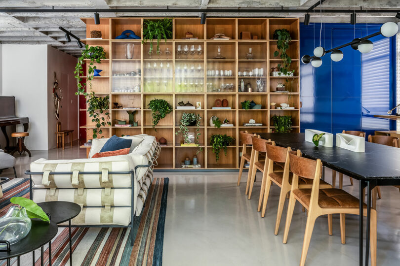 Modern living and dining area featuring a large wooden bookshelf with plants and decorative items. A white sofa, black dining table, and brown chairs are set on a light floor with a striped rug.