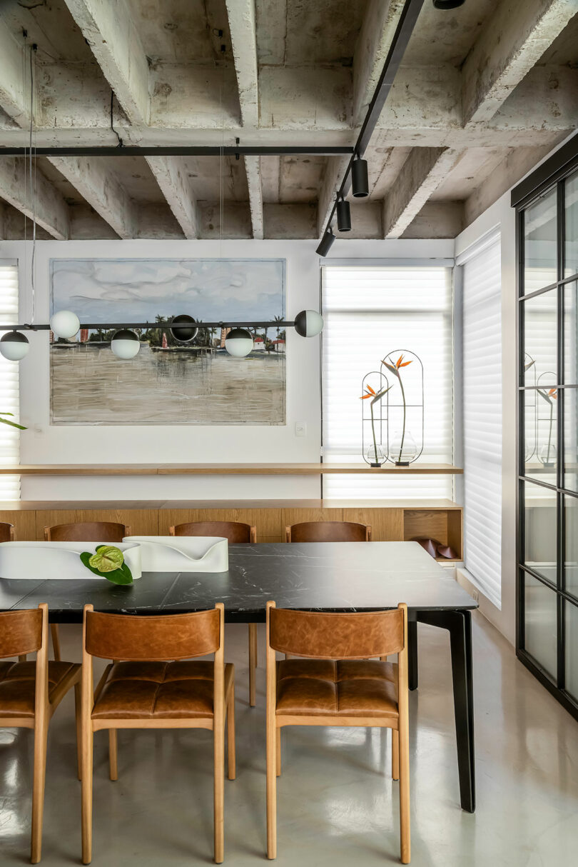 A modern dining room with a black table, six wooden chairs, a large wall painting, and decorative items under exposed concrete beams.