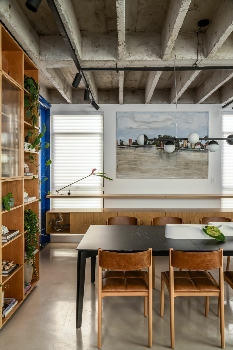 A modern dining room features a black table with four wooden chairs, a large painting on the wall, track lighting, and a bookshelf filled with decor items. The ceiling has exposed concrete beams.