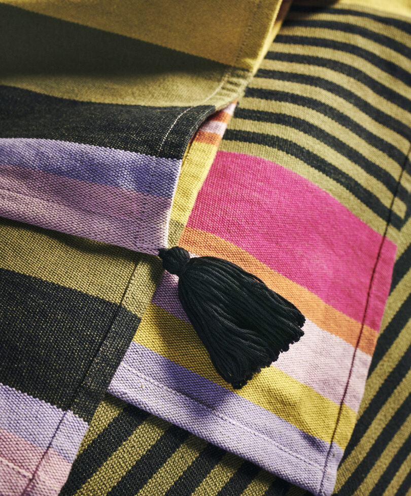 Close-up of a fabric with horizontal stripes in various colors, including black, yellow, pink, and green, featuring a black tassel attachment on one side.