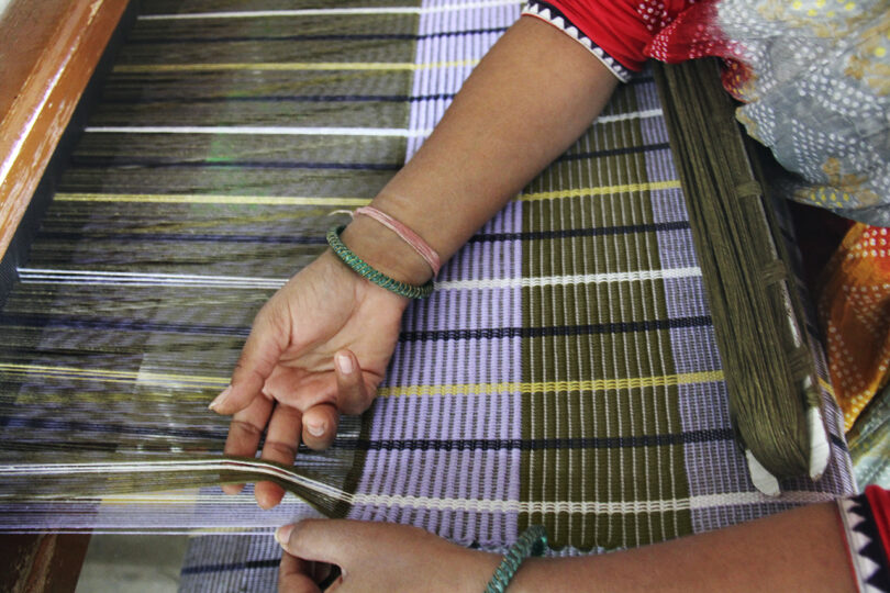 Close-up of a person using a loom to weave a patterned fabric. The hands are manipulating threads, showcasing the intricate weaving process. The person's wrists have simple bracelets.