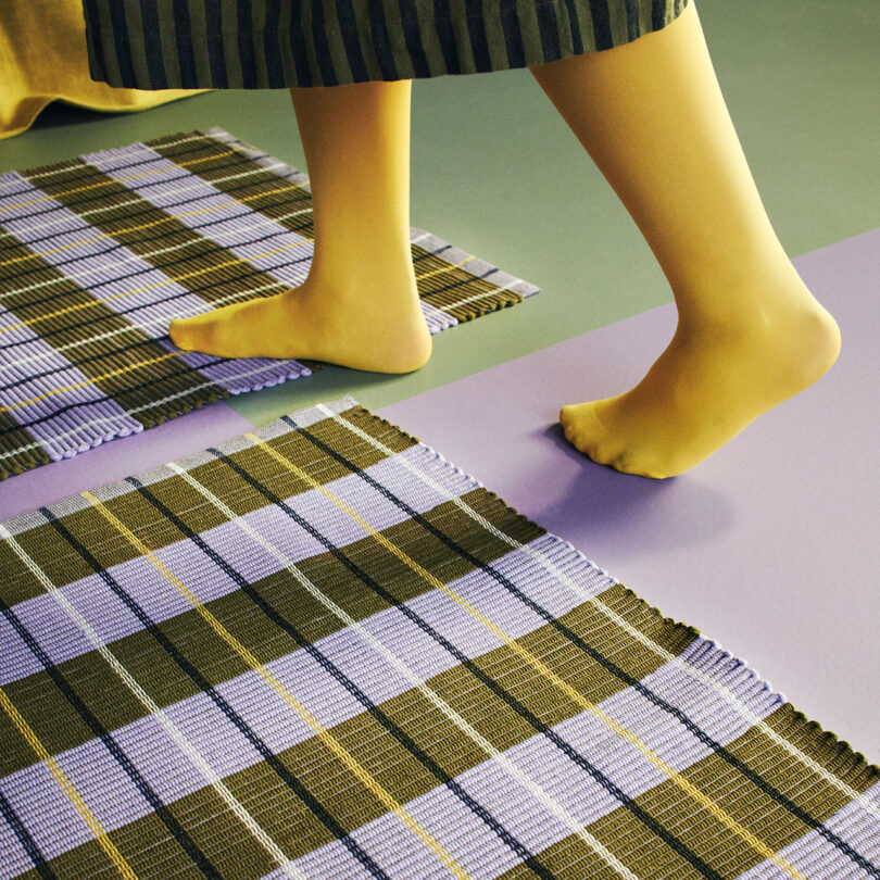 A person walking barefoot on plaid fabric rugs placed on a green and lavender floor. Only the legs and part of a striped dress are visible.