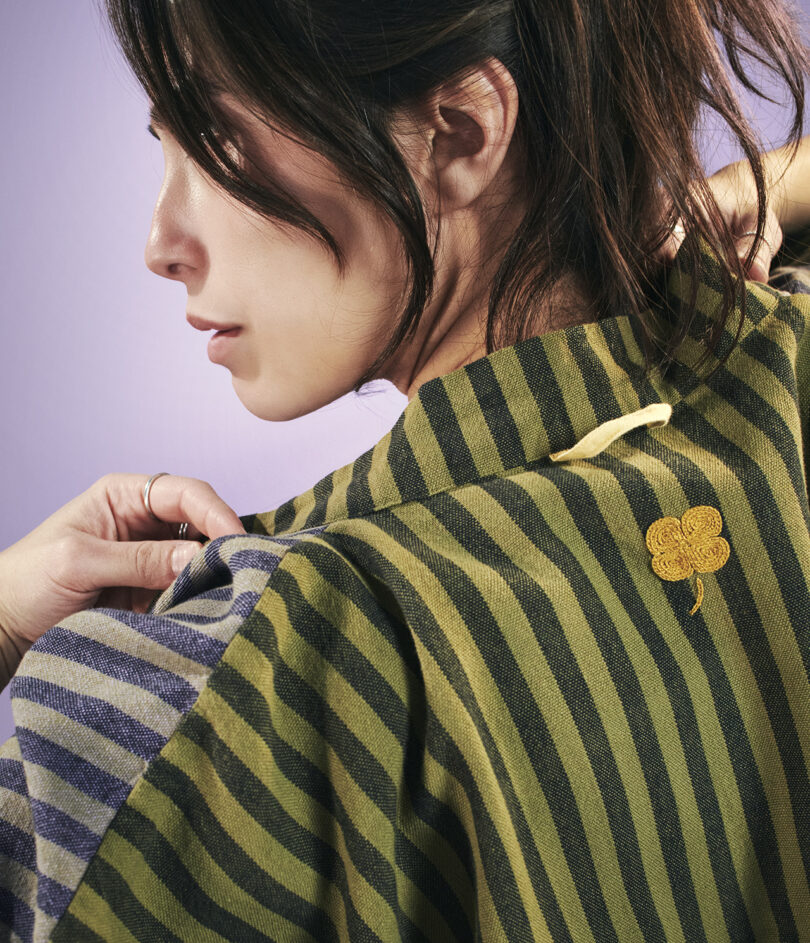 Woman with dark hair wearing a green and black striped garment featuring a small embroidered clover design on the back, adjusting her sleeve.