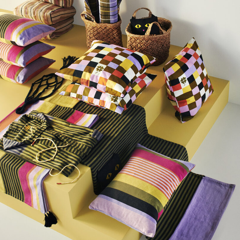 A display of colorful, patterned pillows and textiles arranged on a yellow platform, with baskets filled with folded fabrics in the background.