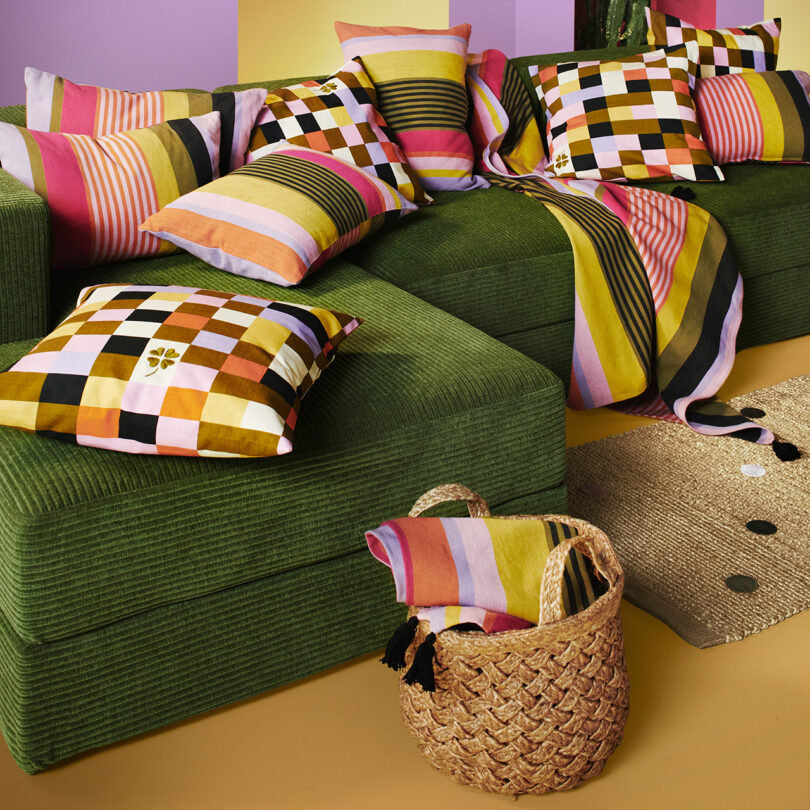 A green sectional sofa covered with colorful, patterned cushions and a matching throw. A woven basket filled with folded textiles sits in the foreground on a jute rug.