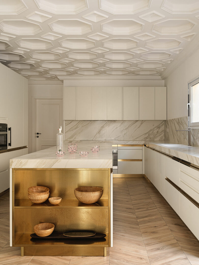 Modern kitchen with light-colored cabinets, marble countertops, and a gold-accented kitchen island displaying wooden bowls. The ceiling, reminiscent of a Marina Braginskaya design, features an intricate geometric pattern.