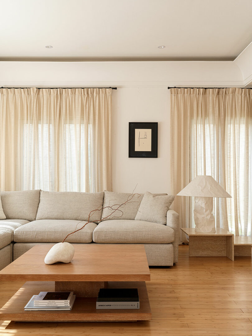 A minimalist living room features a light beige sectional sofa, a wooden coffee table with books, and a table lamp on a side table. Cream curtains cover the windows, and a framed artwork by Marina Braginskaya graces the wall.