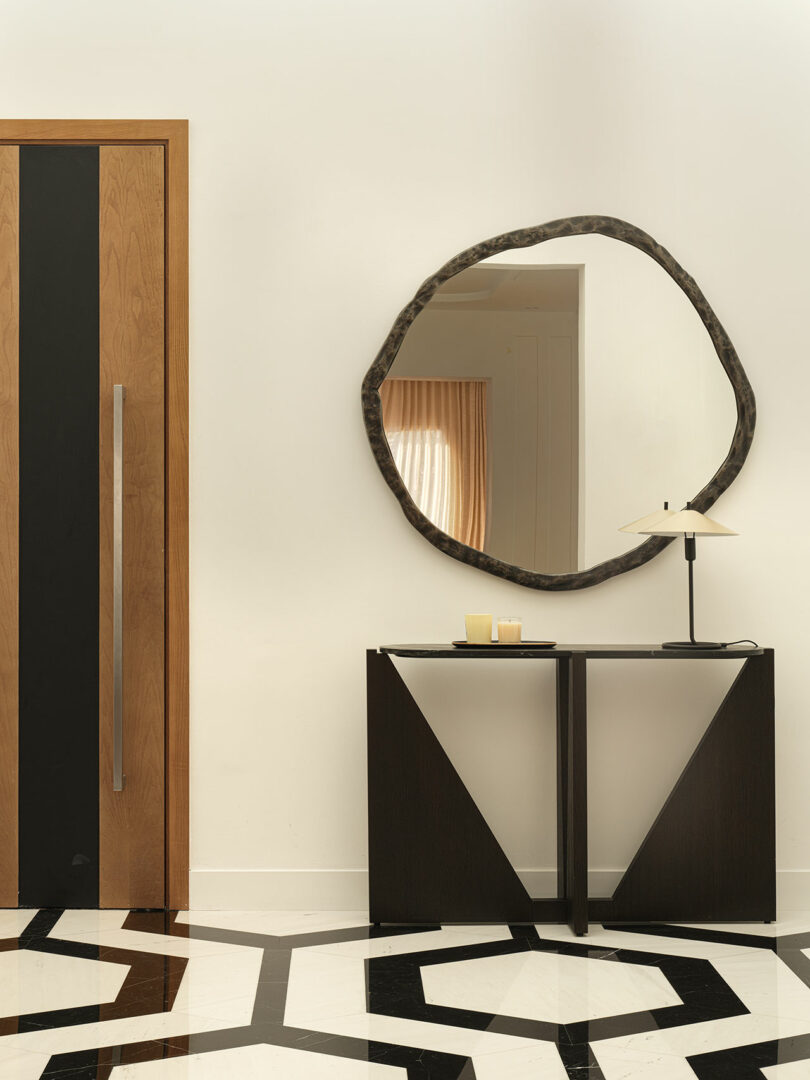 Modern hallway with a geometric-patterned floor, a Marina Baisel-designed wooden door, circular mirror, black console table, two candles, and a sleek lamp.