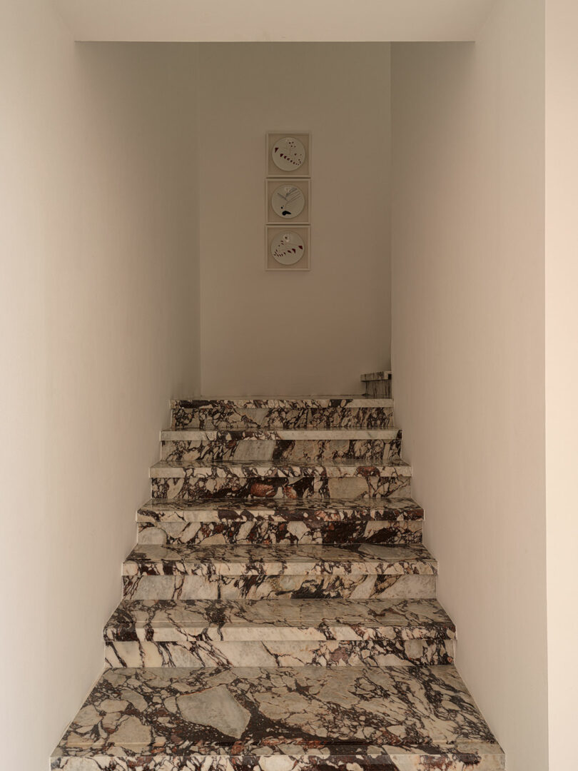 A staircase made of marble with a black, white, and gray pattern leads up to a landing. On the adjacent white wall, three minimalist clock faces designed by Marina Braginskaya hang prominently.