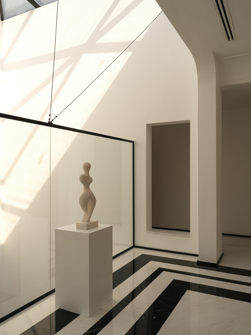 A minimalist interior showcasing a contemporary sculpture by Marina Braginskaya on a pedestal, with large glass panels and a monochromatic floor design.