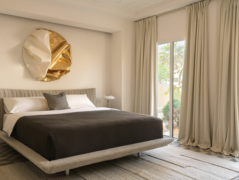 Modern bedroom with a low platform bed adorned with black and white bedding, a bedside table, and sculptural wall art inspired by Marina Braginskaya. Floor-to-ceiling curtains cover a large window overlooking greenery.