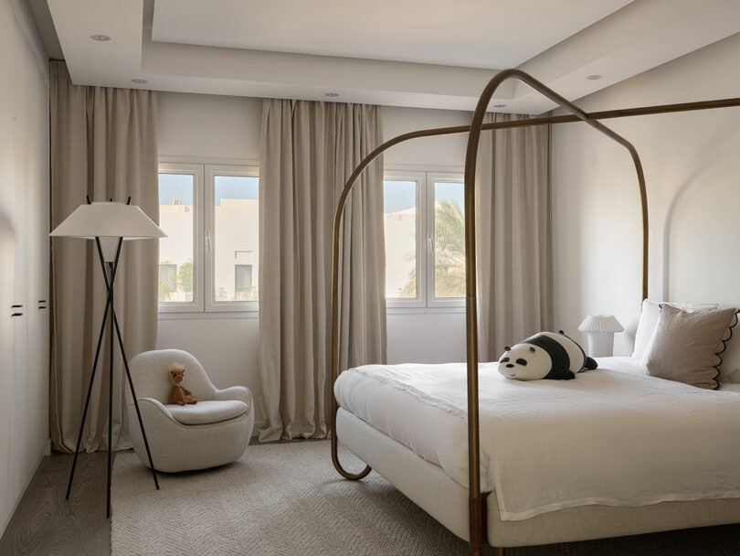 A minimalist bedroom featuring a canopy bed with white bedding, a teddy bear on a chair, and a panda pillow on the bed. Natural light filters through windows adorned with beige curtains, reminiscent of a design by Marina Braginskaya.