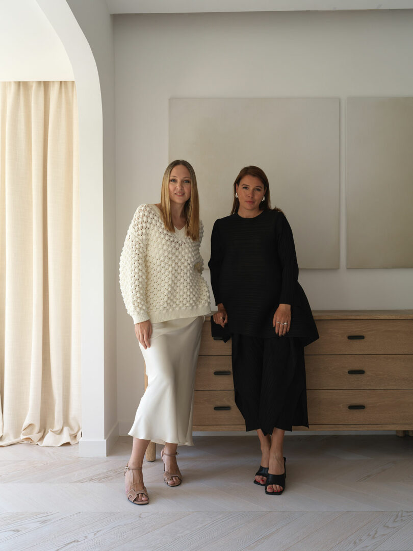 Two women, Marina Baisel and Marina Braginskaya, stand side by side in a minimalist room with light-colored walls and wooden furniture. One wears a white outfit, the other a black one. They both look towards the camera.