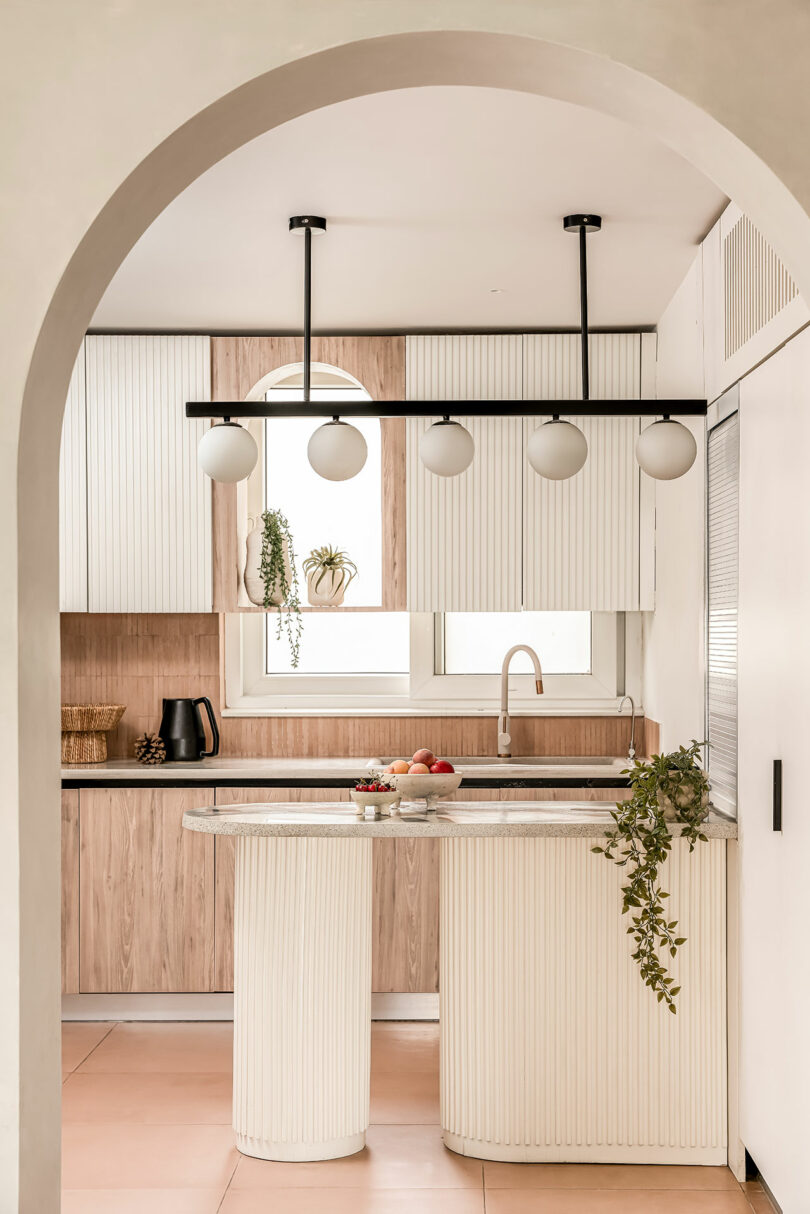 Modern kitchen with an archway entrance, featuring light wood and white cabinetry, a central island with plants, contemporary pendant lighting, and a window above the sink.