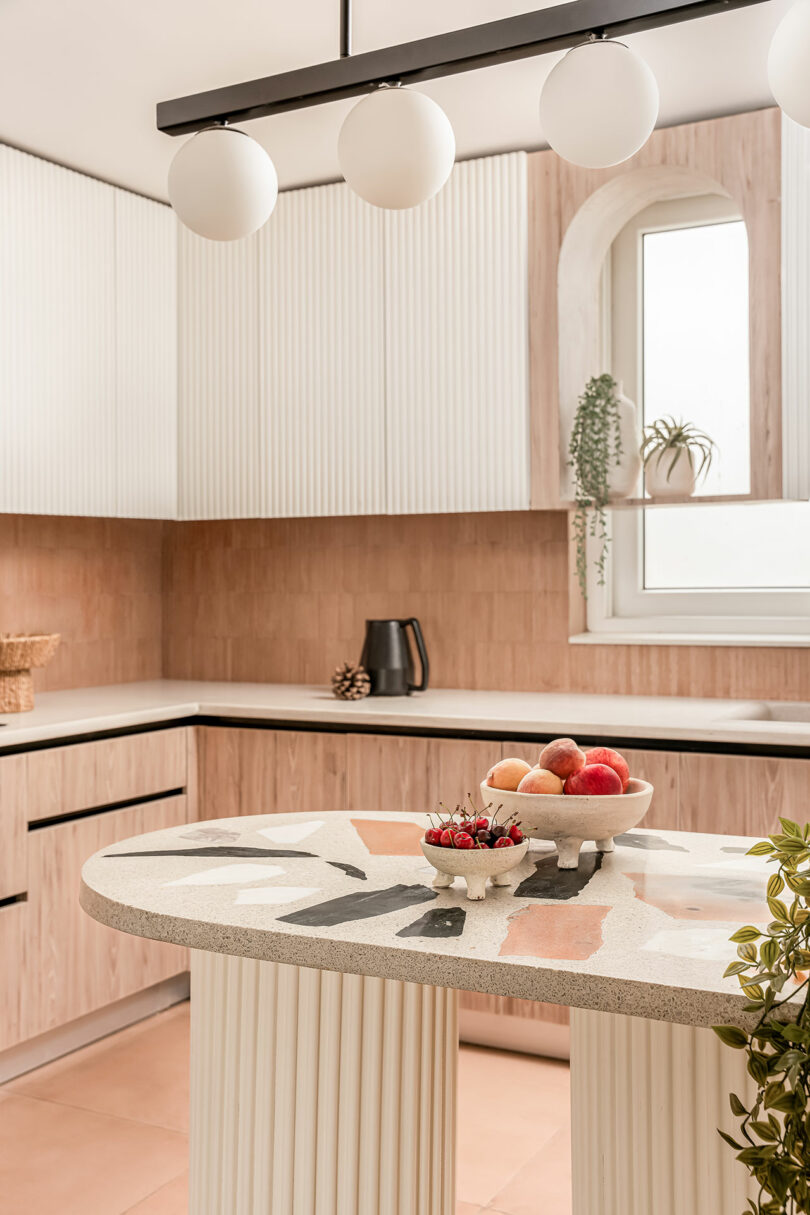 Modern kitchen with light wood cabinets, a patterned island, a black kettle, a window, and bowls of fruit and berries. The room is decorated with plants and has a minimalist, clean design.