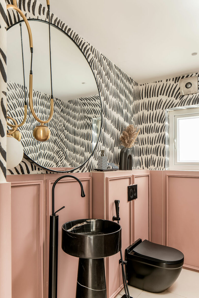 A modern bathroom featuring a black sink, black toilet, large round mirror, and decorative wallpaper with a pink panel accent. A small window with natural light is visible on the right.