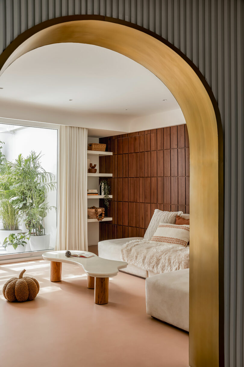 Living room with modern decor featuring a curved archway, white sofa, rustic wooden paneling on the wall, plants by a large window, and a pumpkin-shaped ottoman on the floor.