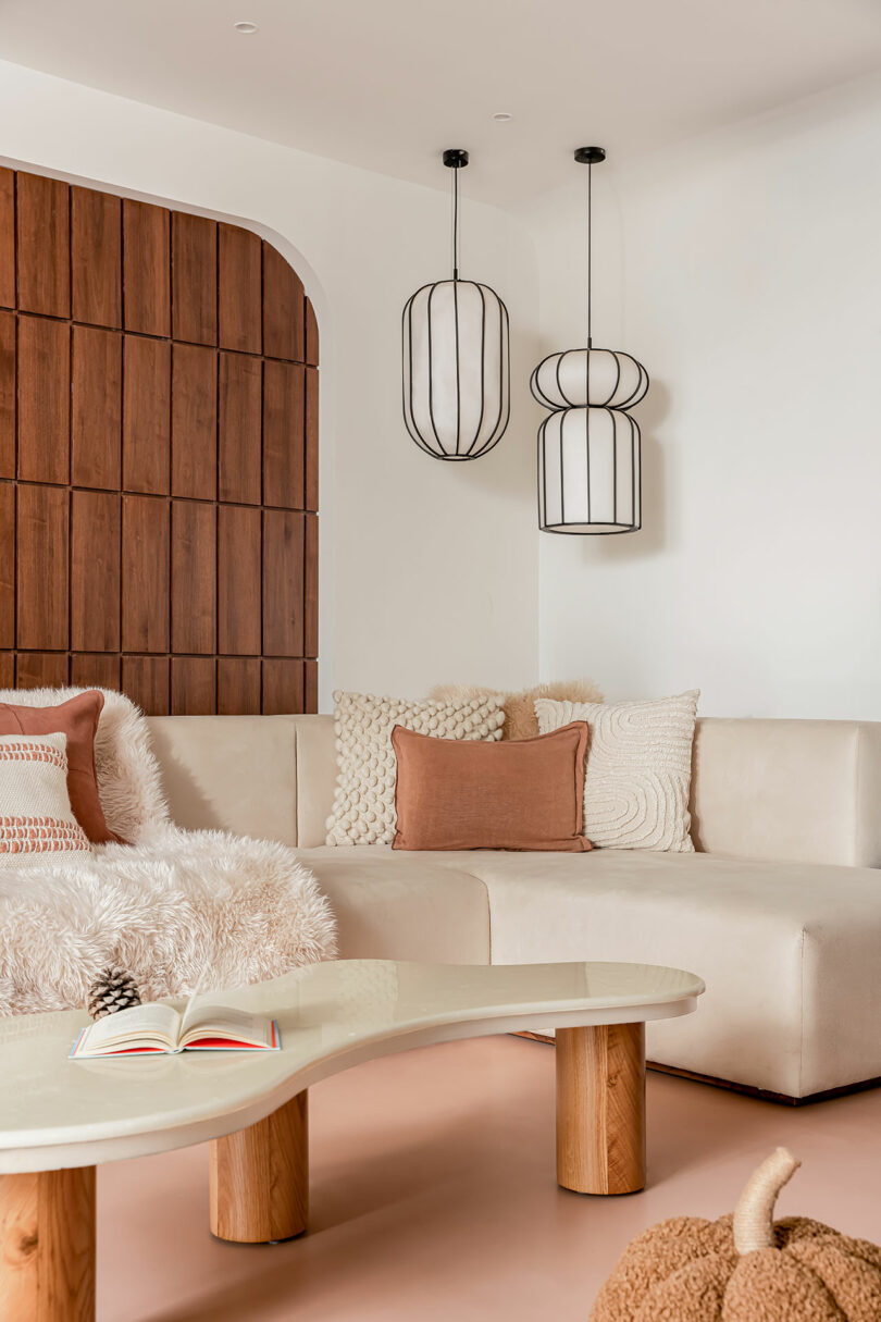 A modern living room with a white sectional sofa, various textured pillows, hanging light fixtures, a wooden accent wall, and a glass coffee table with light wood legs. An open book lies on the table.