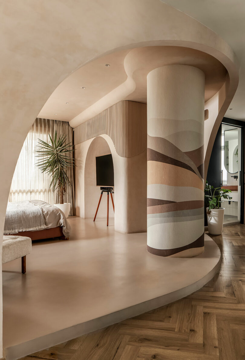 Modern bedroom with a curved architectural design, featuring a central pillar with artistic patterns, neutral tones, a large plant, and a wall-mounted TV.